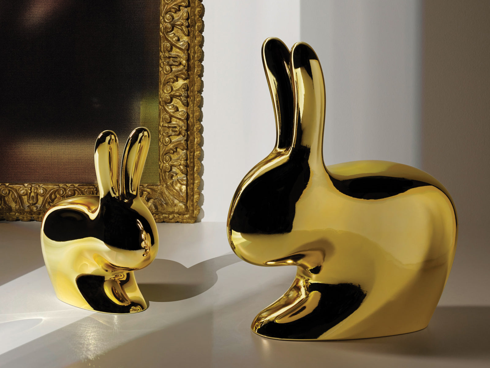 Two copper Rabbit Chairs, a large and a small, on a white surface beside an ornate framed artwork.