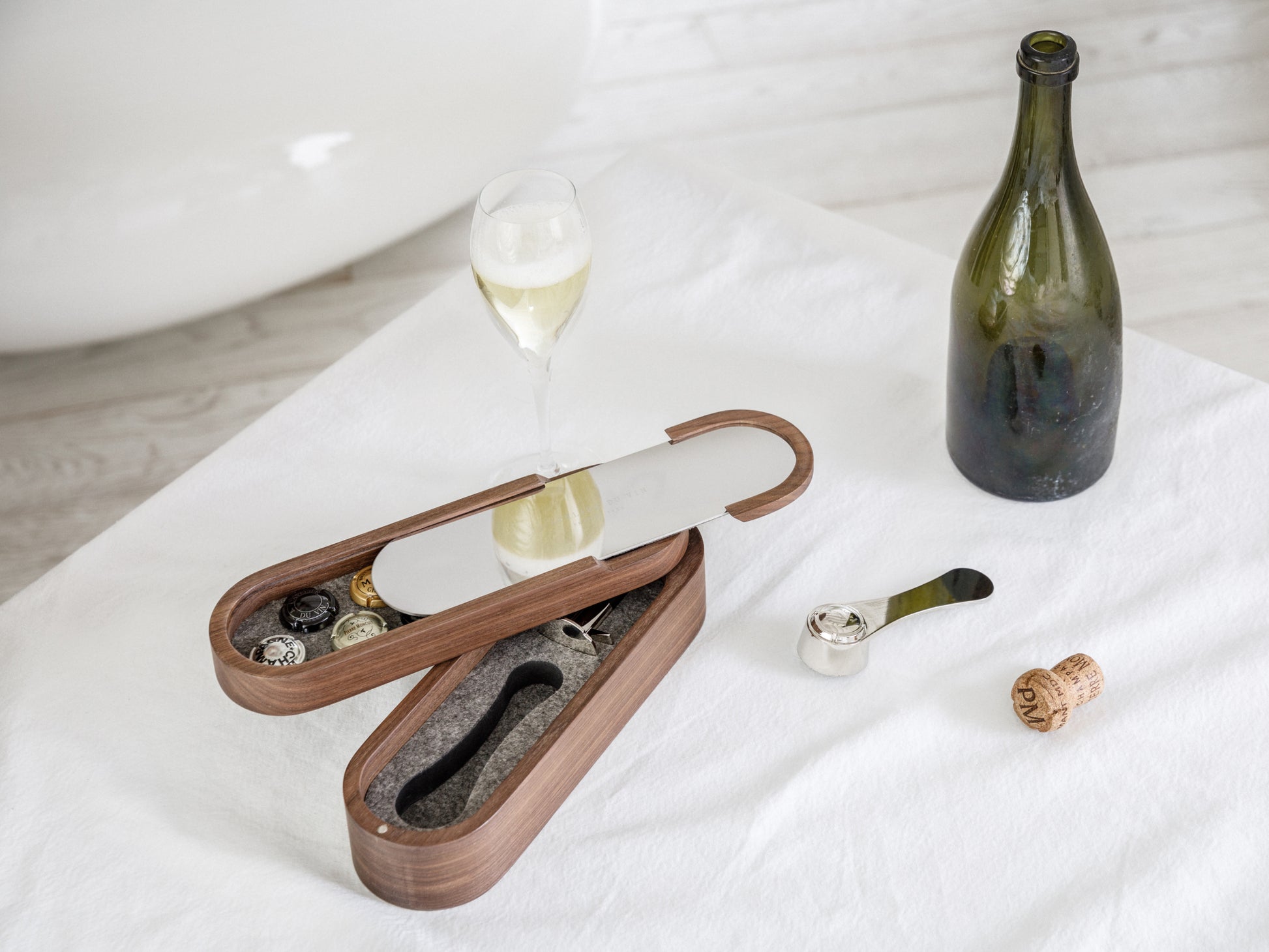 Opened walnut Sparkling Set with champagne glass and bottle on a white surface beside the open box showing its interior compartments.