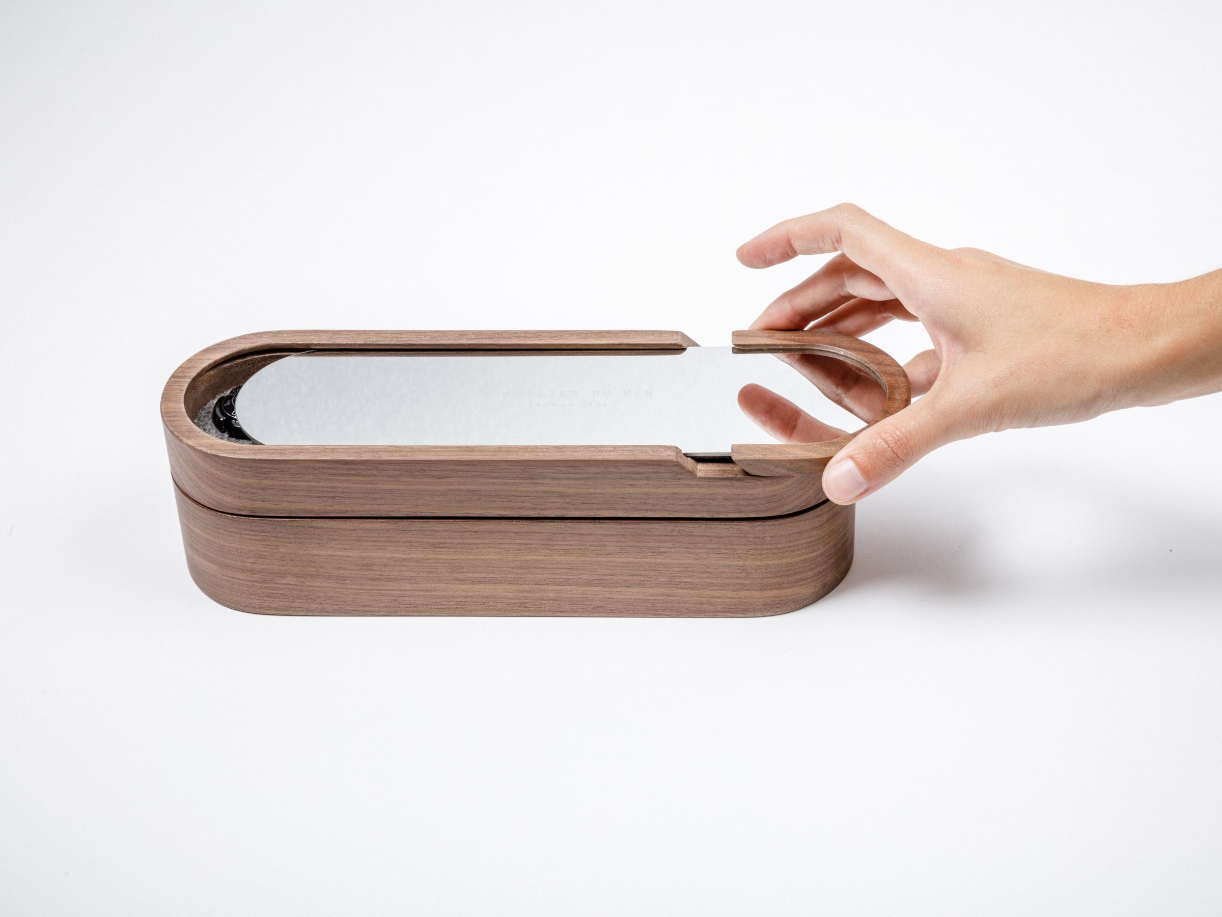 Hand lifts the curved walnut lid of the Sparkling Set, revealing the slim reflective metal panel.