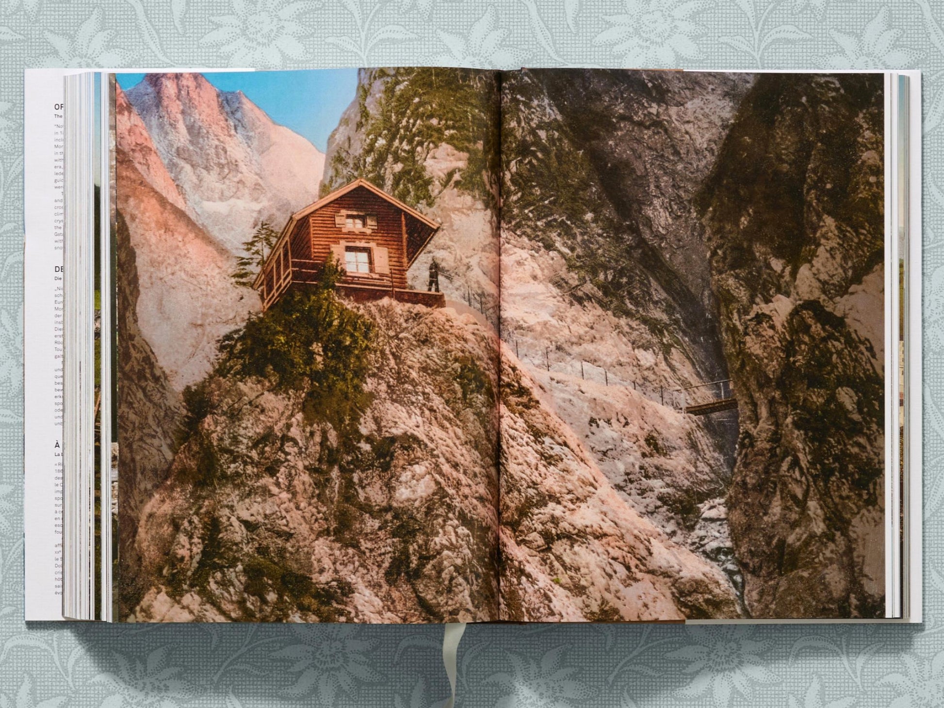 Wooden alpine hut perched on a rocky slope with a narrow bridge spanning a gorge.