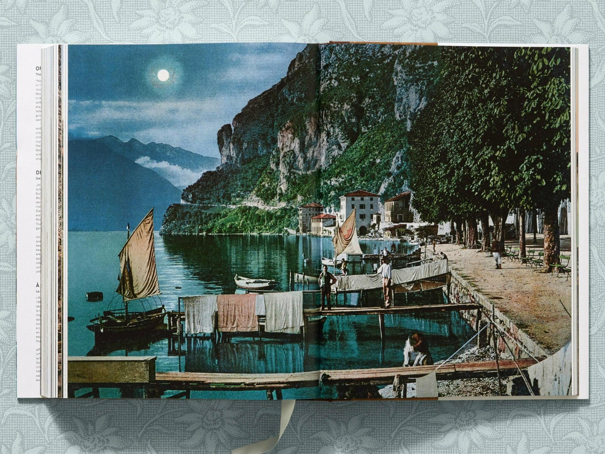 Lakeside alpine village beside a rocky cliff with sailboats and people by a dock and a white house