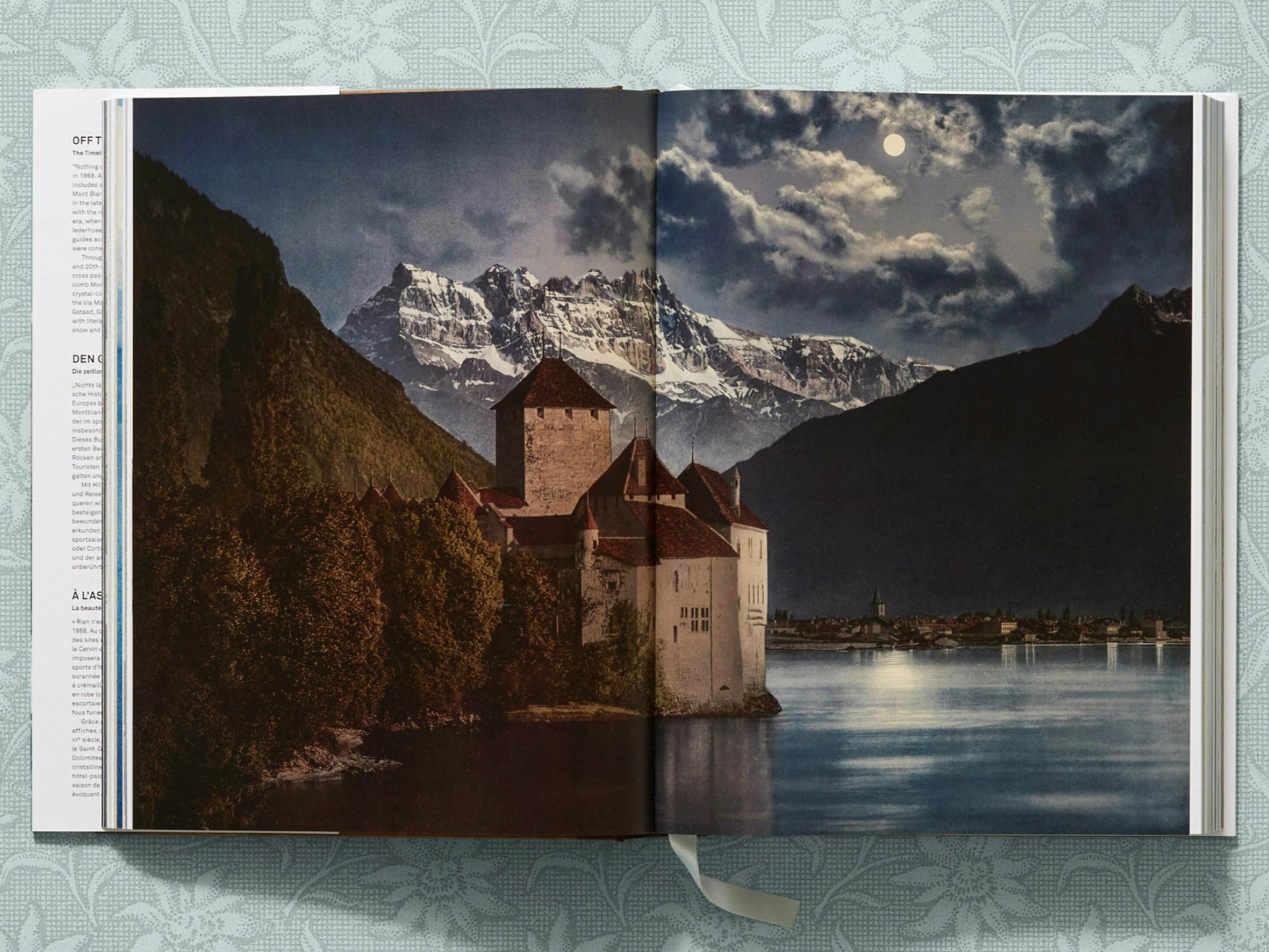 Castle perched by a lake with dramatic snowcapped mountains and cloudy sky in the background.