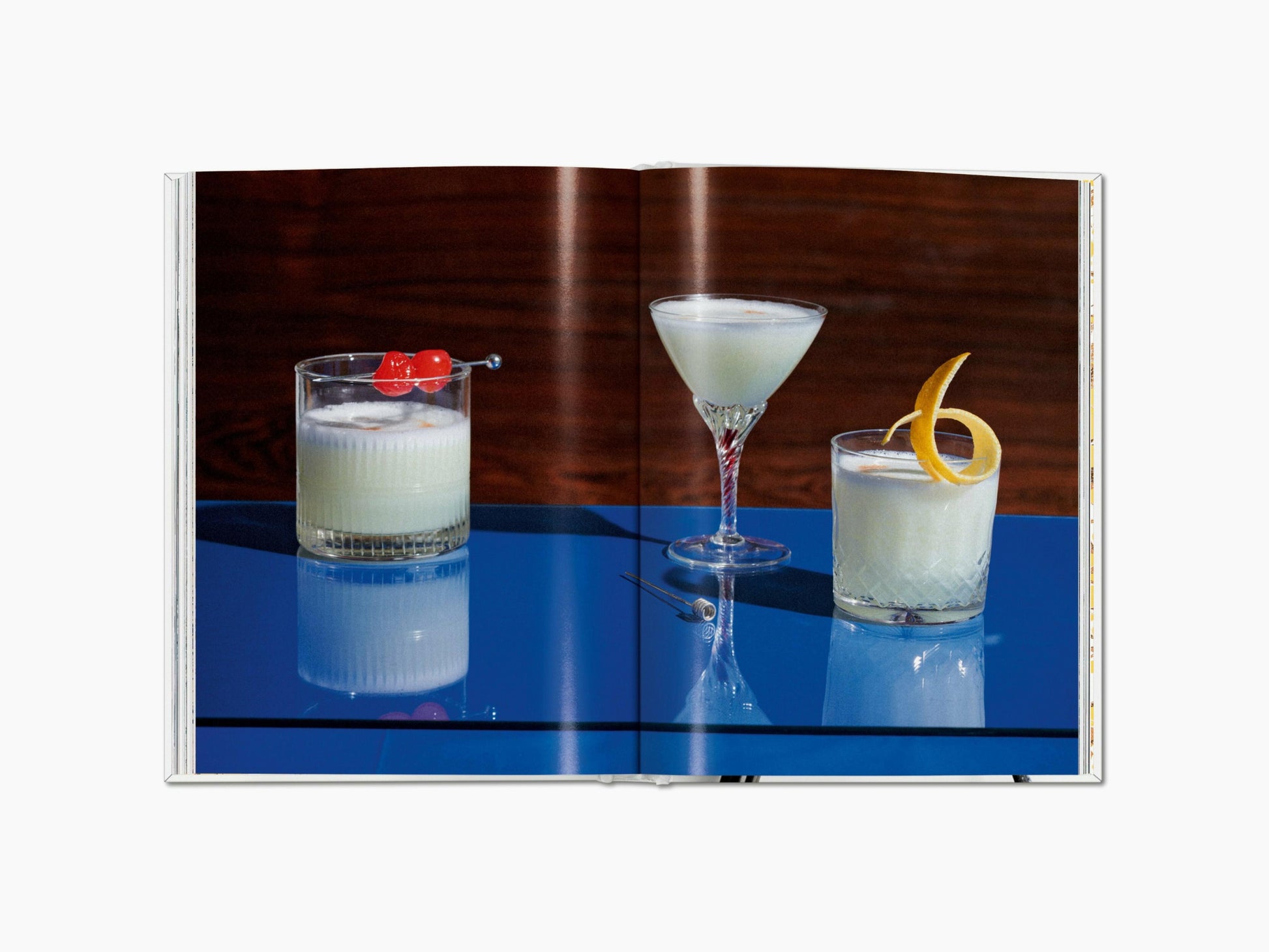Three cocktails on a glossy blue bar: left a rocks glass with cherry garnish, middle a stemmed martini, right a short glass with a lemon twist.