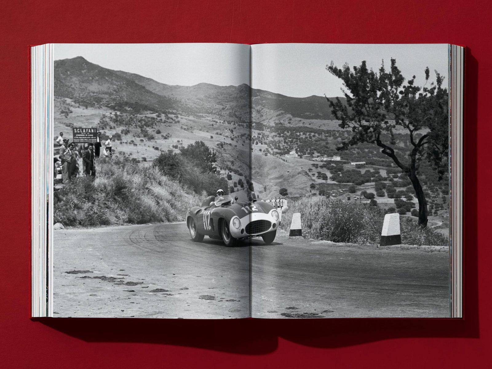 Open book spread showing a black and white photo of a vintage race car rounding a mountain road, with spectators on the hill and a tree on the right.