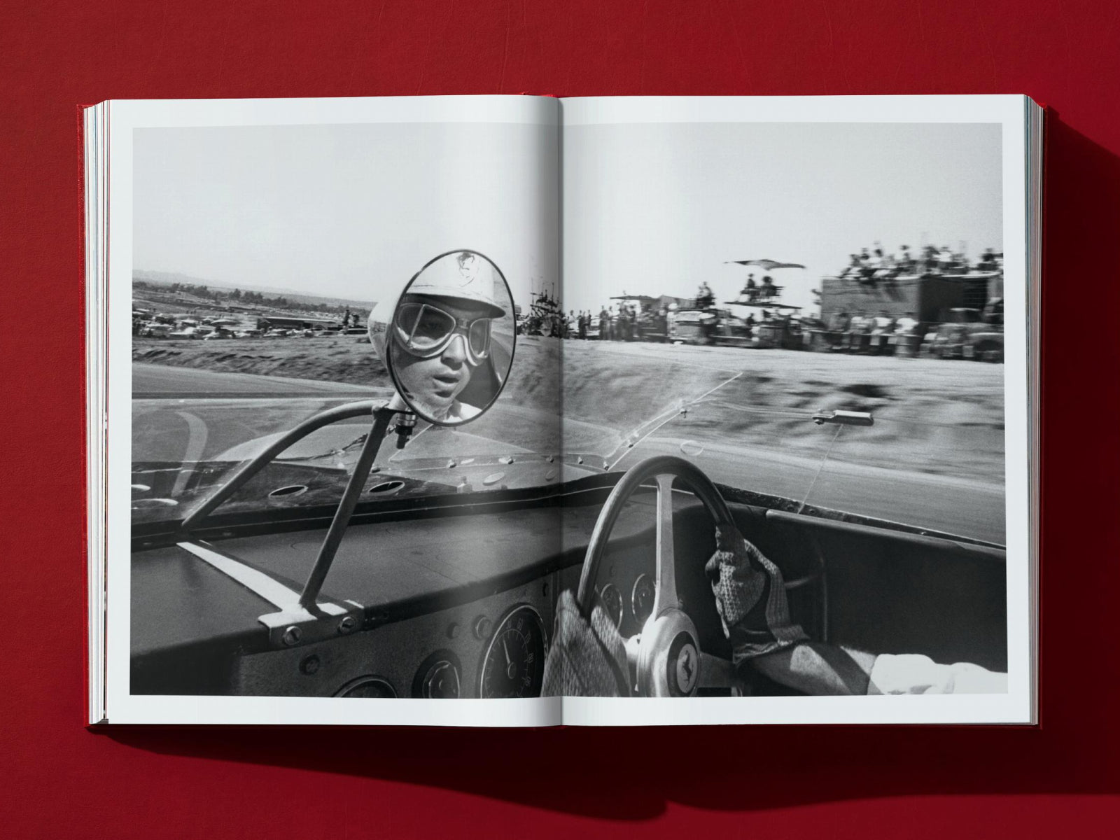 Driver wearing goggles in a vintage race car cockpit with the track and crowd visible through the windshield.