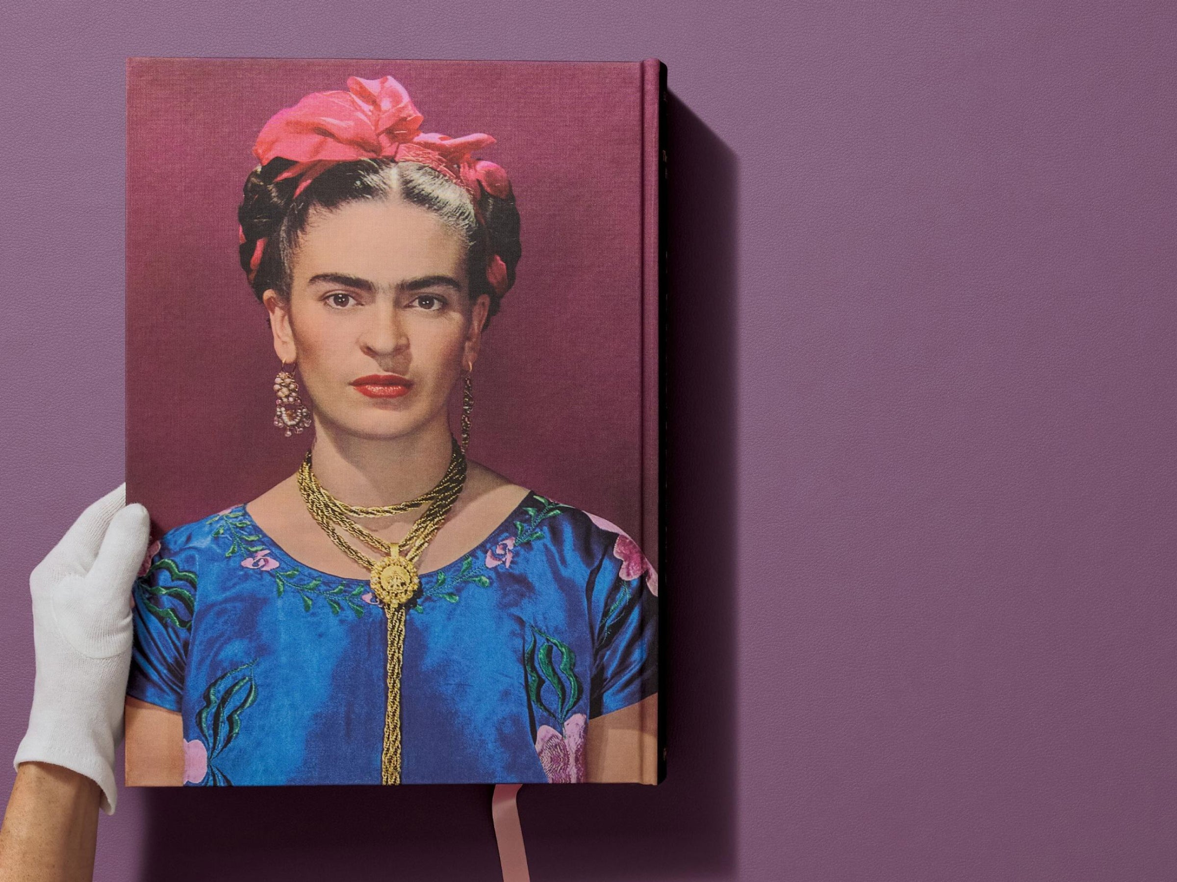 Portrait of Frida Kahlo on the burgundy cover of Frida Kahlo The Complete Paintings, held by a white gloved hand.