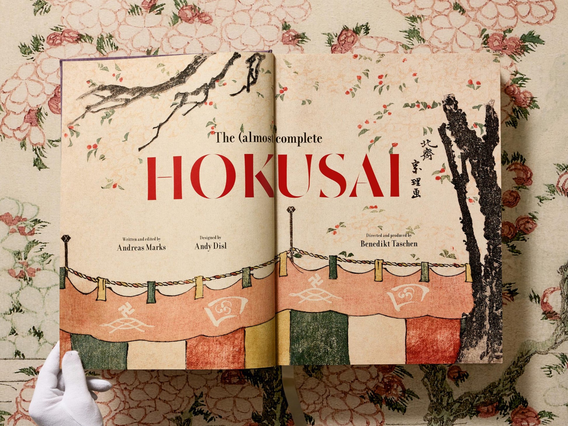 Open Hokusai book spread showing bold red HOKUSAI on the left and an illustration on the right held by a gloved hand.