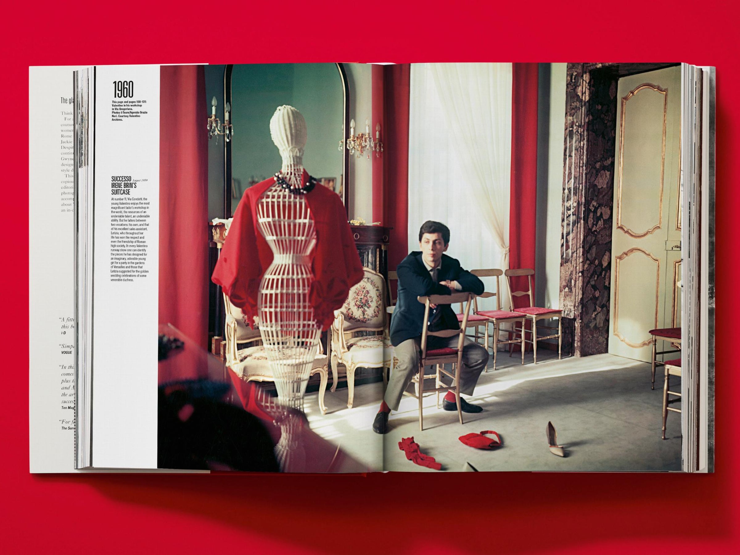 Color photograph of a fashion studio with a man seated among chairs, a red garment on a dress form, and red drapery.