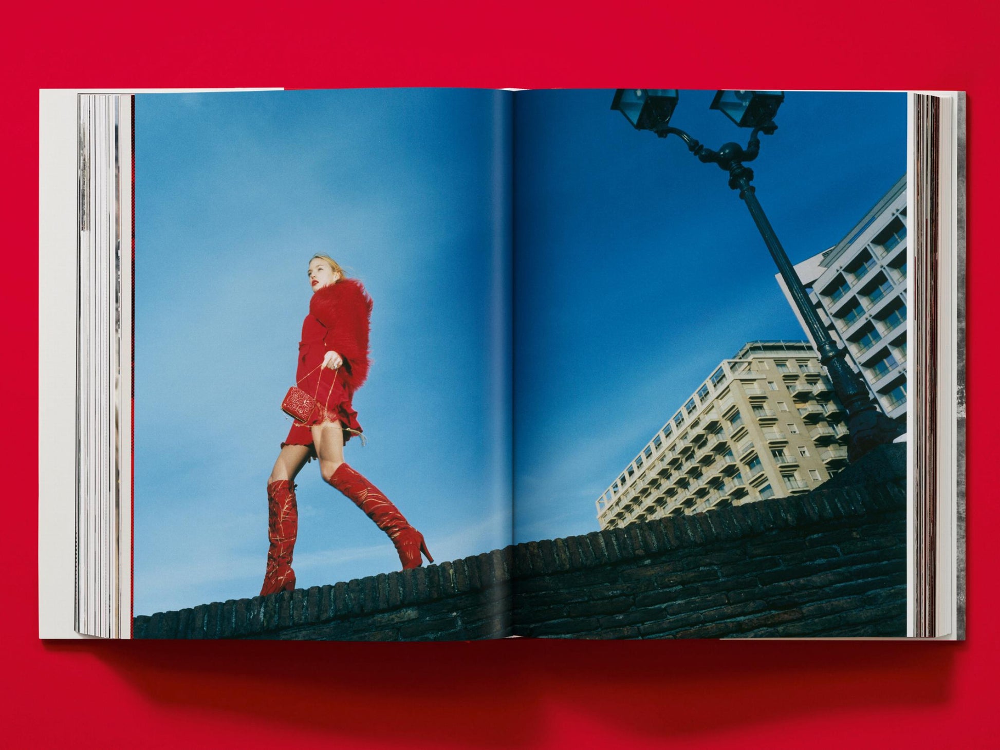 Model in red fur coat and red boots stands atop a brick wall against a bright blue sky.