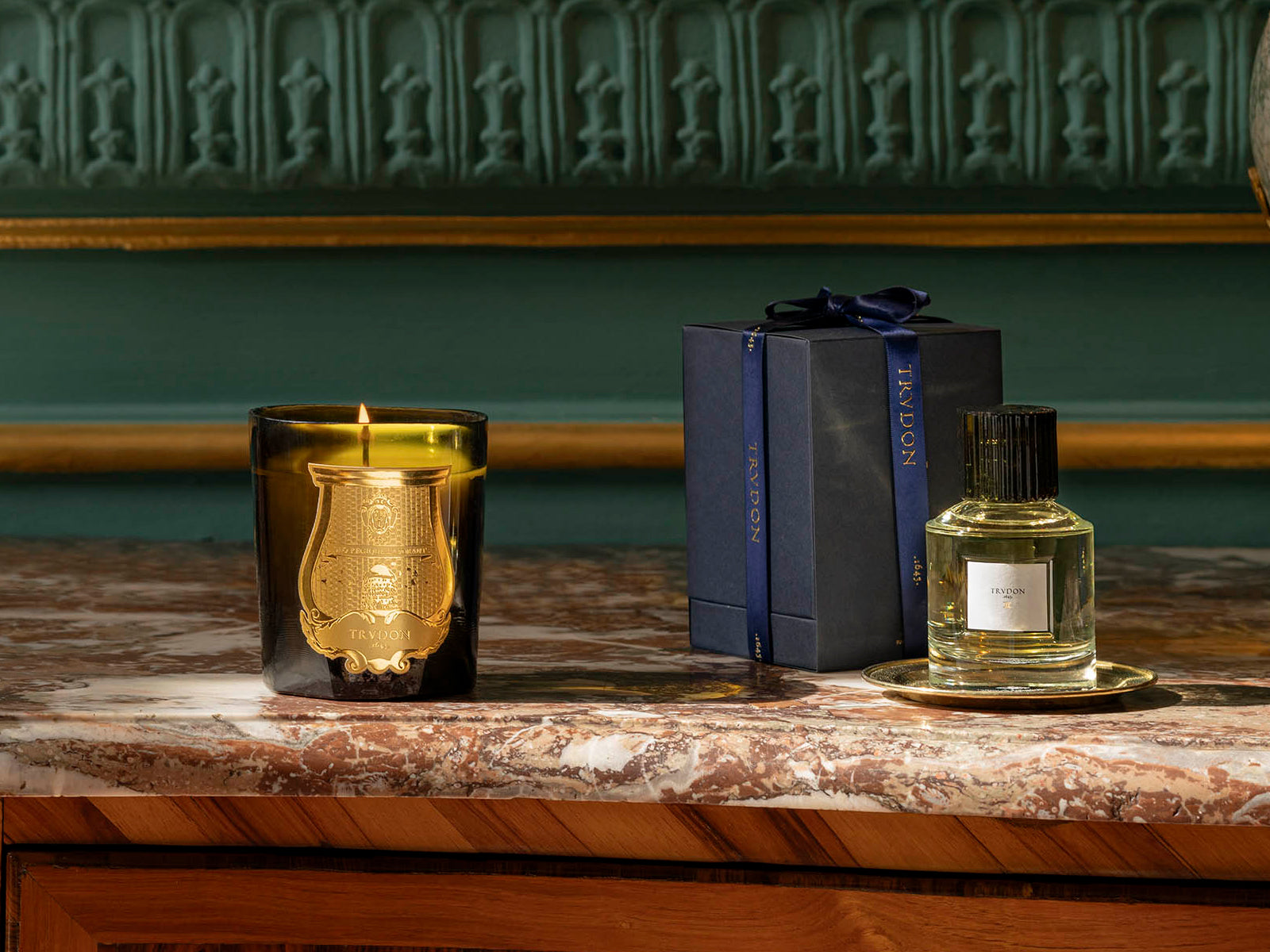 Joséphine Floral Garden candle in dark glass with gold crest beside a navy gift box tied with a ribbon and a perfume bottle on a marble counter.