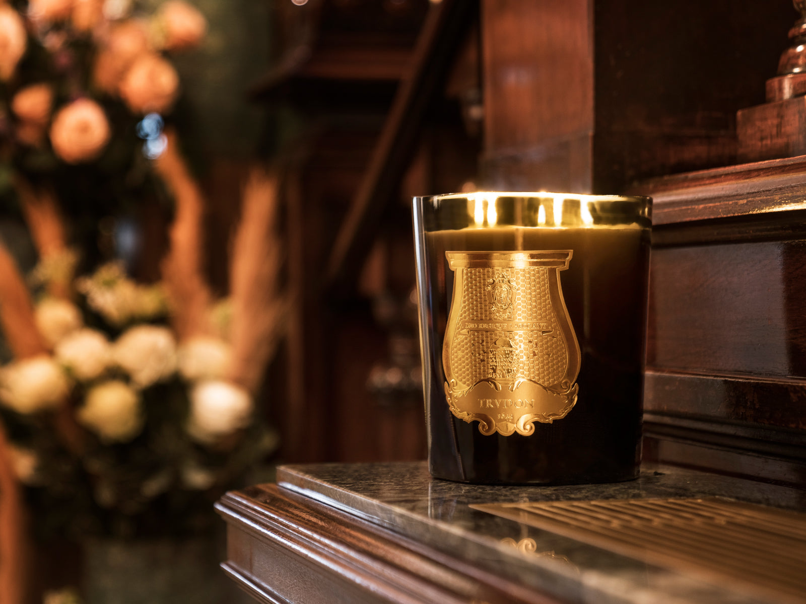 Joséphine Floral Garden candle in a frosted white glass with a gold crest on a warm wooden surface with a blurred floral backdrop