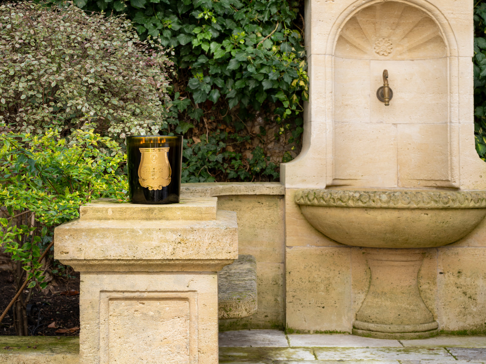 Grande Abd El Kader Moroccan Mint Tea candle in a dark glass container on a stone pedestal beside a fountain.