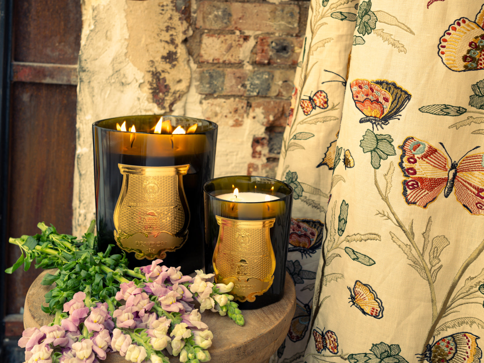 Grande Abd El Kader Moroccan Mint Tea candles in dark glass with gold crests, two lit beside a floral curtain.