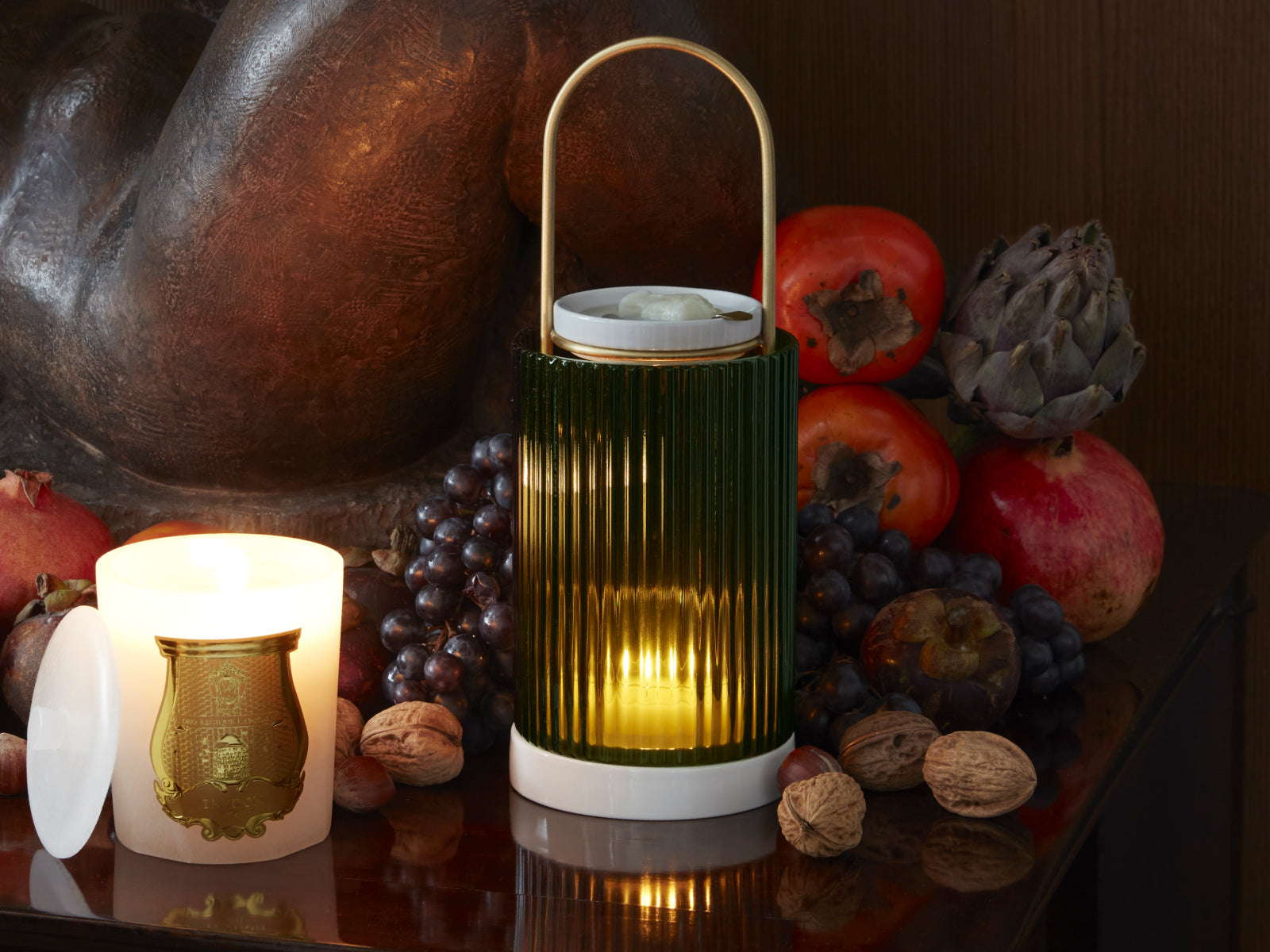 Green ribbed glass lantern with a gold handle and a warm candle glow beside fruit and nuts.