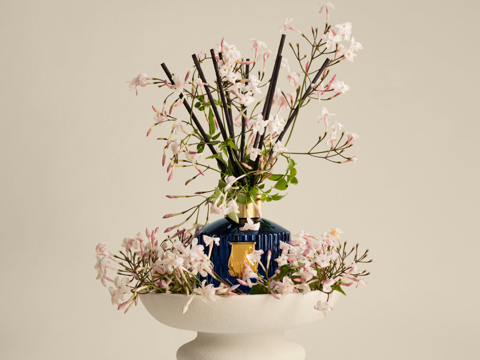 Blue glass diffuser bottle with a gold emblem among pink jasmine blossoms arranged on a white pedestal