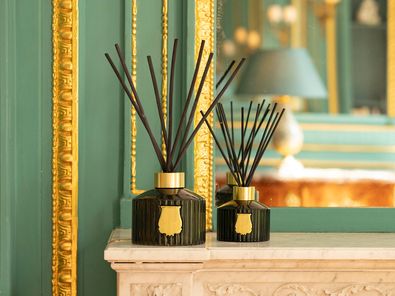 Two Grand Diffusers with tall black reeds and gold collars on a marble mantel against emerald green walls with ornate gold trim