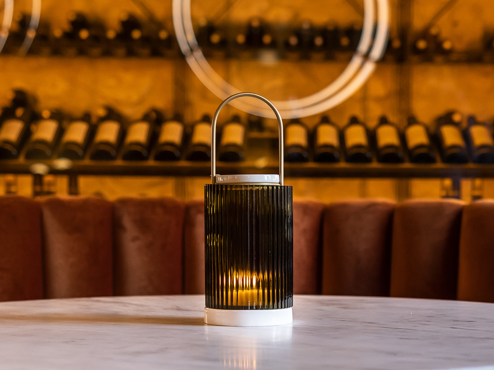 La Promeneuse diffuser with ribbed black body white base and curved handle glowing on a marble table in a warmly lit bar.