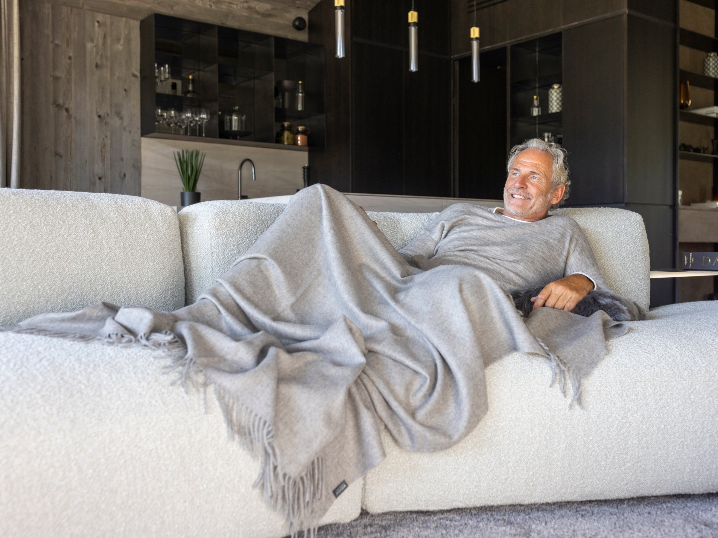 A man lounging on a light sofa wrapped in a gray alpaca plaid blanket in a modern open living space.