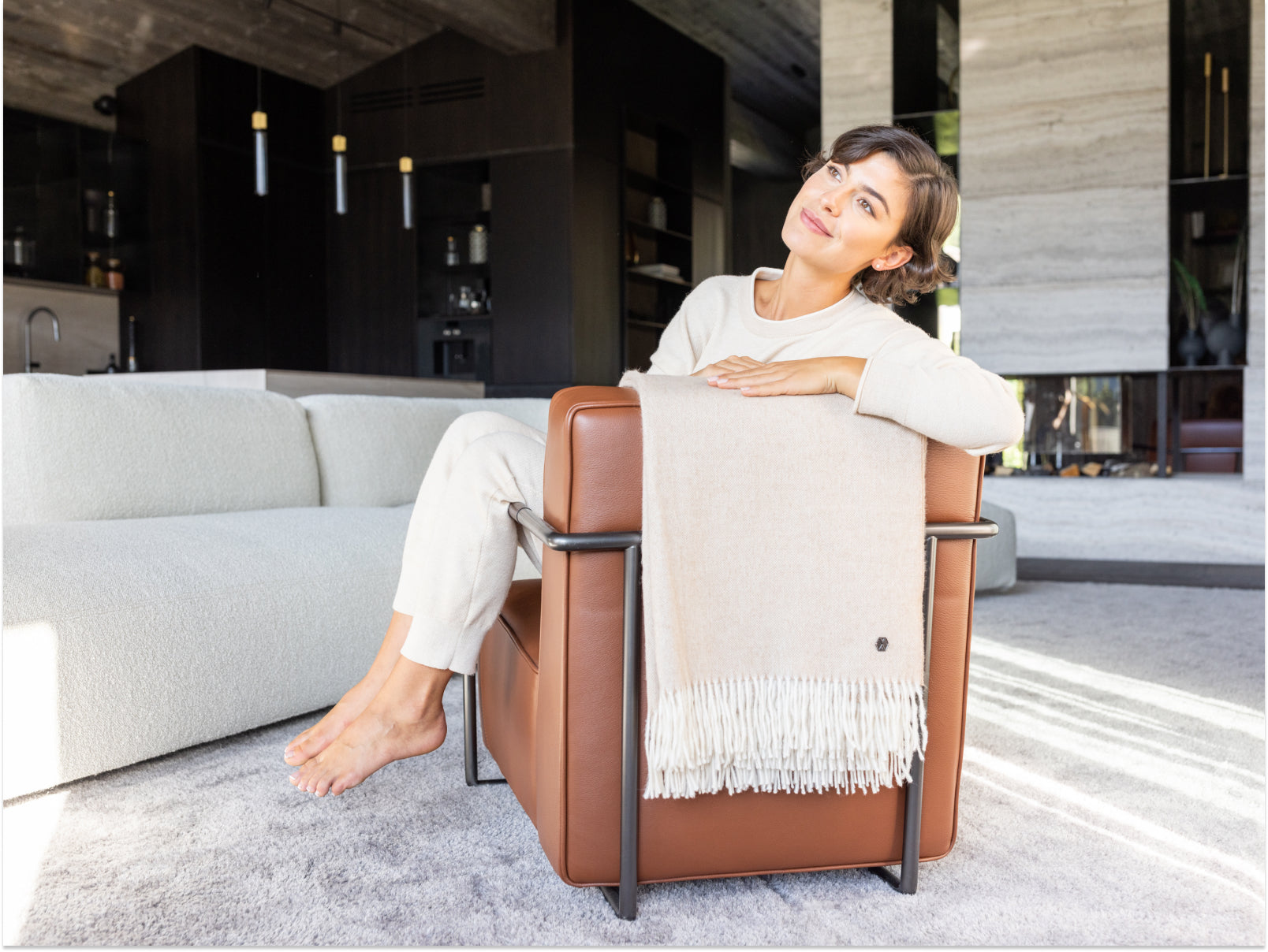 Woman lounging on a brown leather chair with a beige alpaca plaid throw draped over the back in a modern living room.