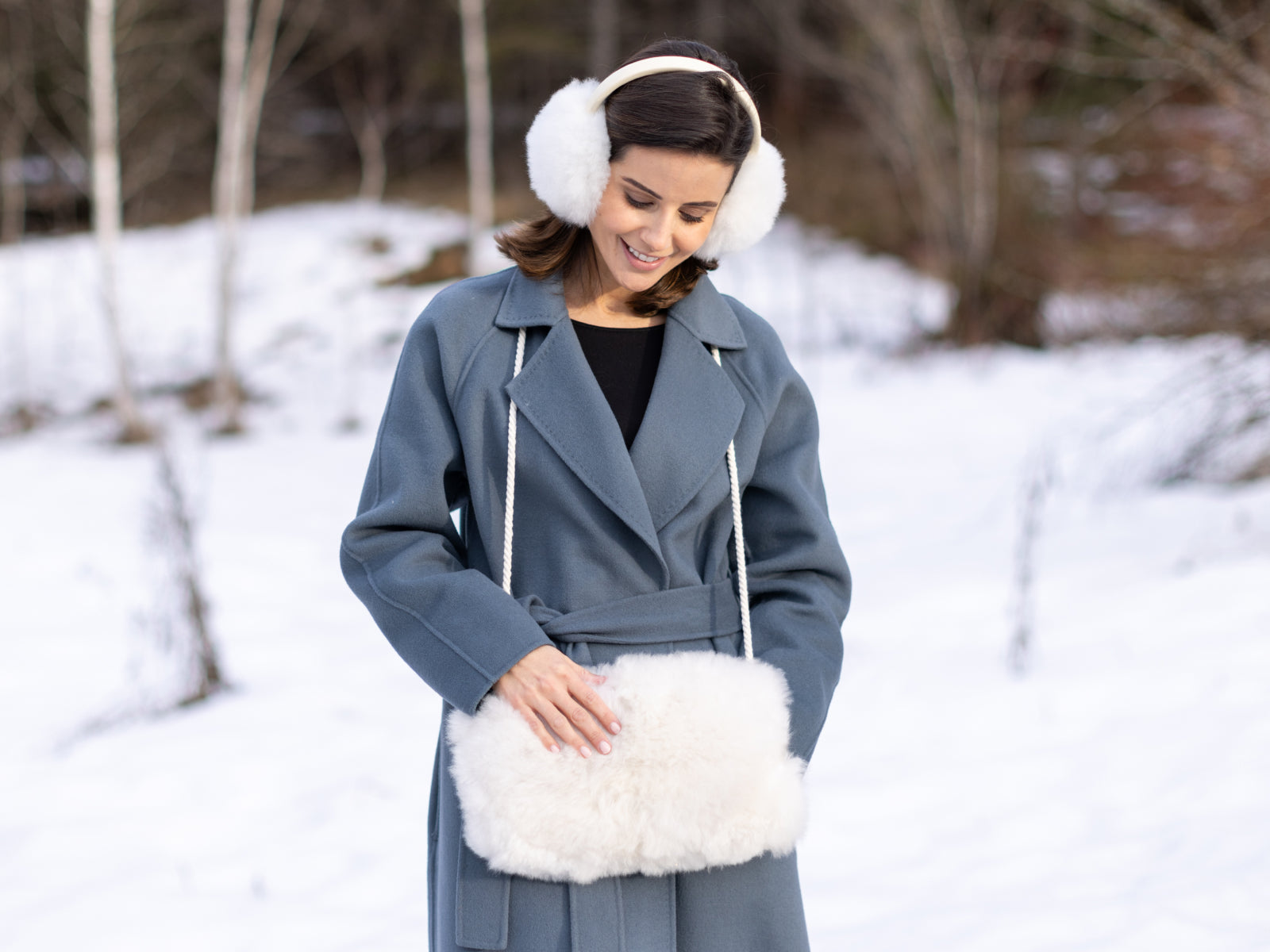 Woman in a blue coat outdoors in the snow wearing white alpaca earmuffs and holding a fluffy pocket muff.