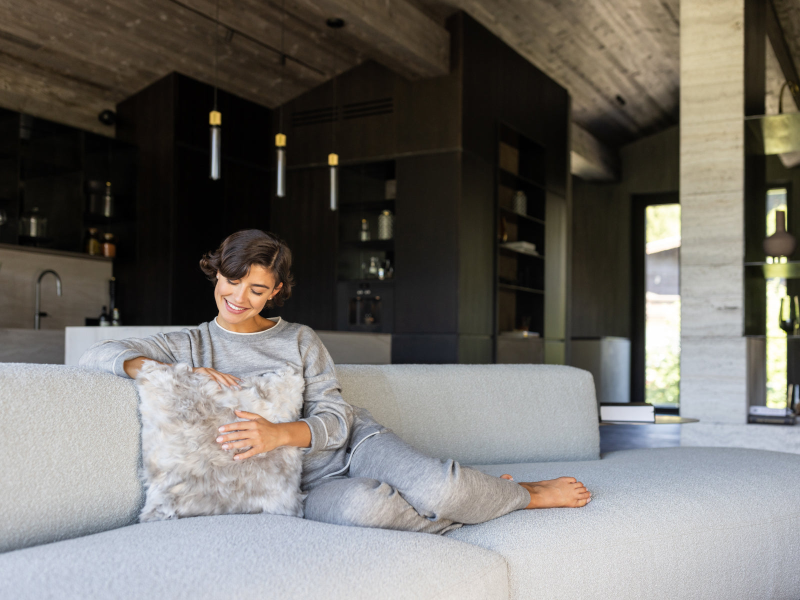 Smiling woman lounging on a light gray sofa, cuddling a plush alpaca fibre cushion.