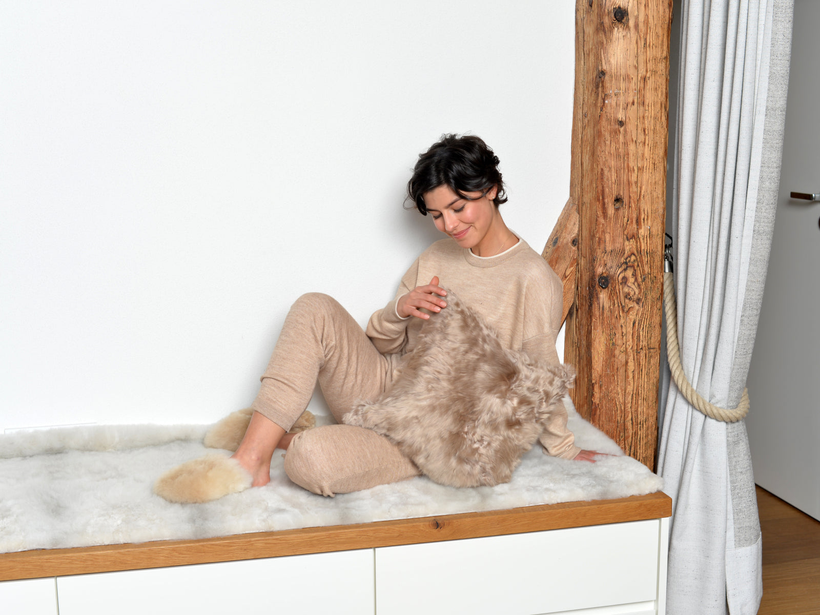 Beige-clad woman sits on a white fur surface by a wooden beam, holding a plush alpaca fibre cushion.