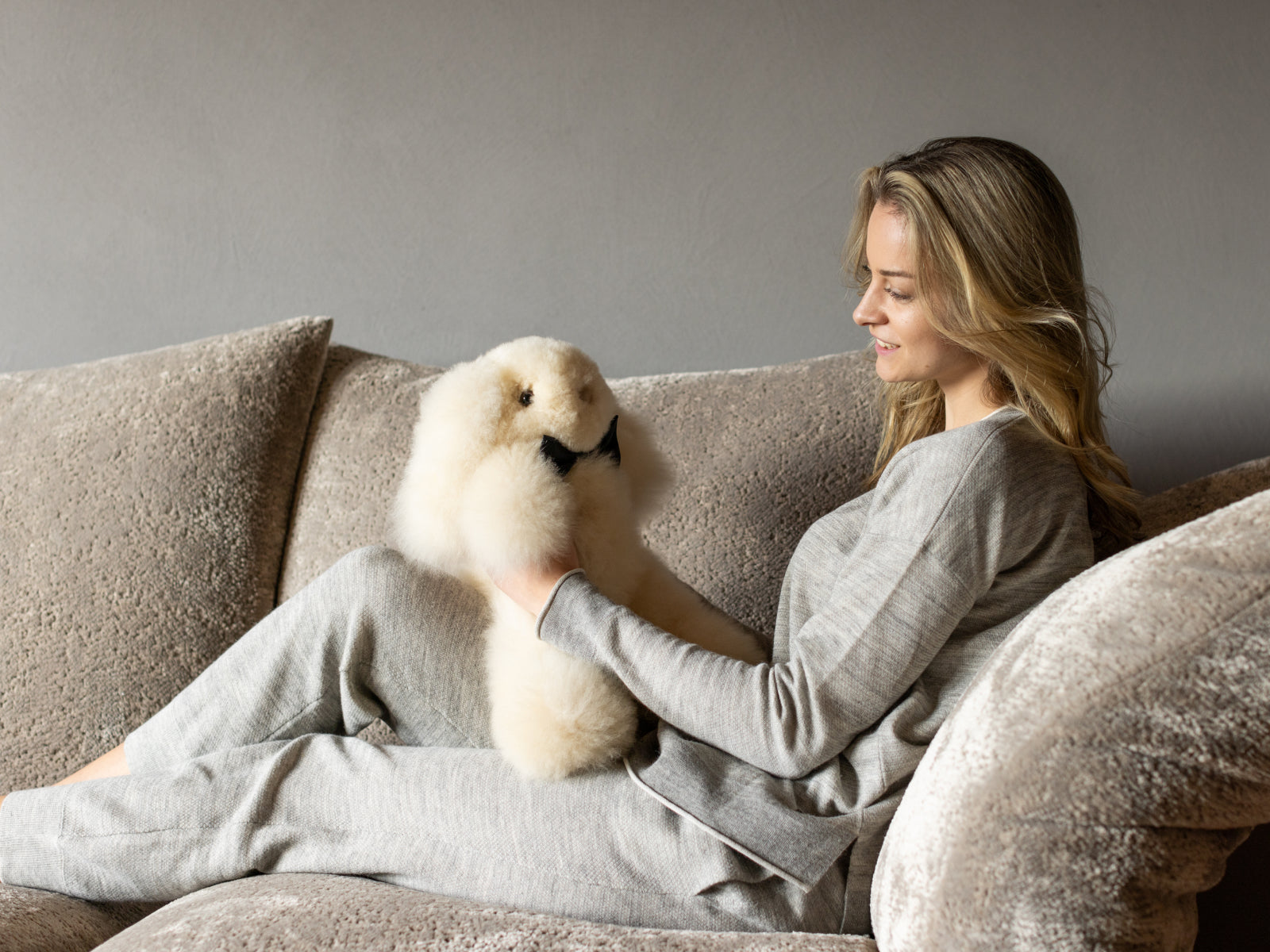 Smiling woman on a beige sofa hugs Milo the cream alpaca plush rabbit with a black bow tie.