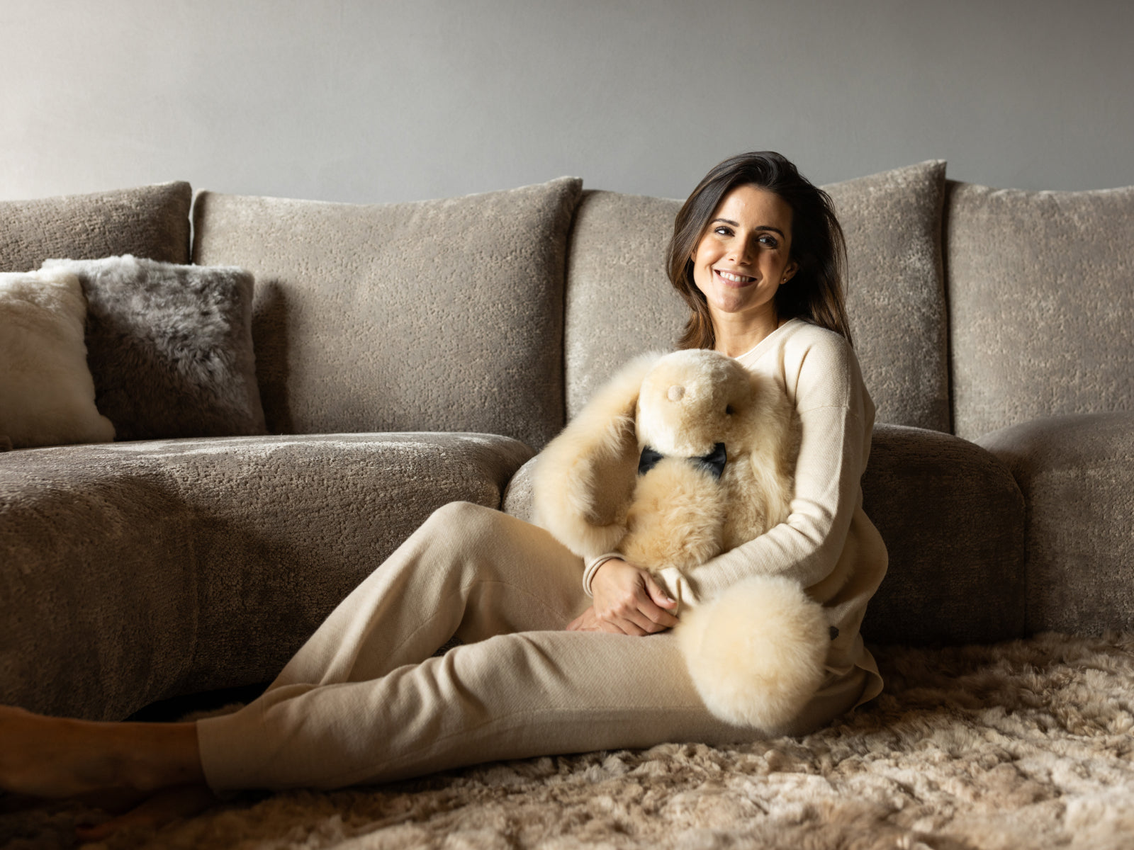 Woman in a beige sweater sits on the floor cuddling Milo the fluffy alpaca plush rabbit.