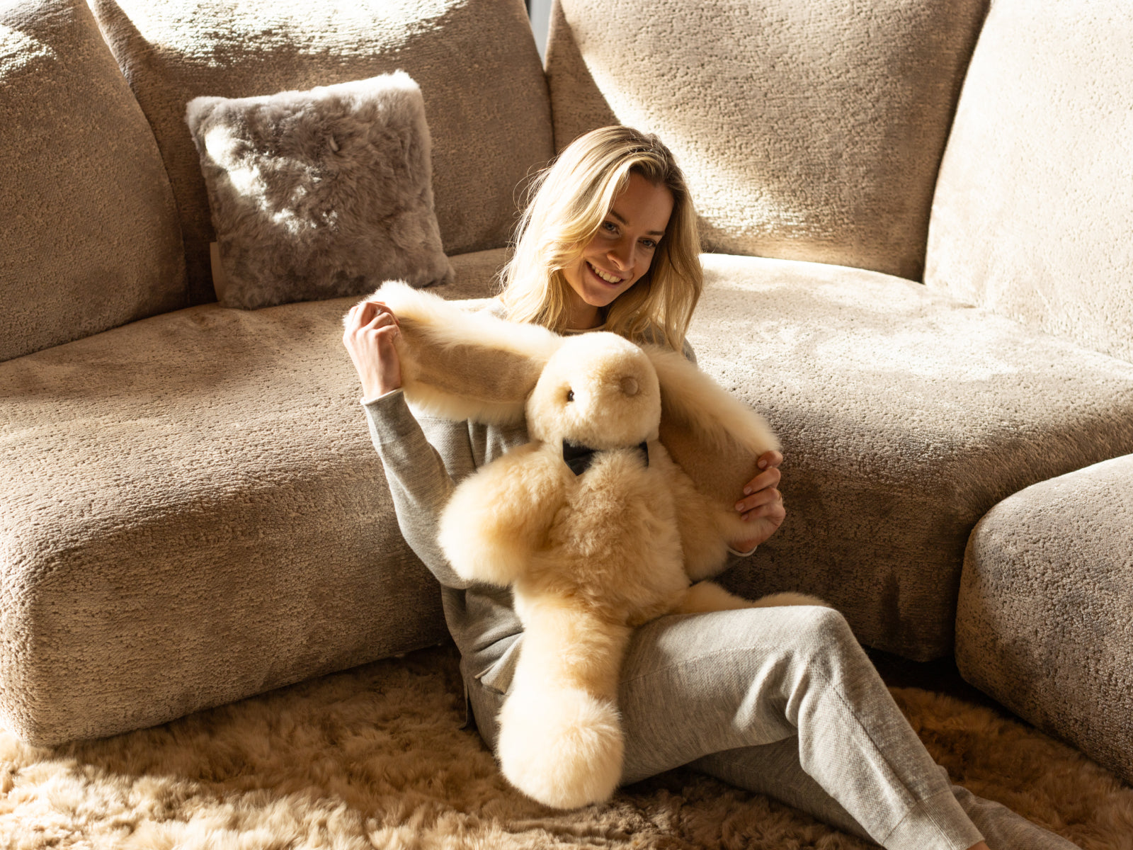 Woman in grey loungewear sits on a beige sofa and cuddles Milo the fluffy alpaca plush rabbit.