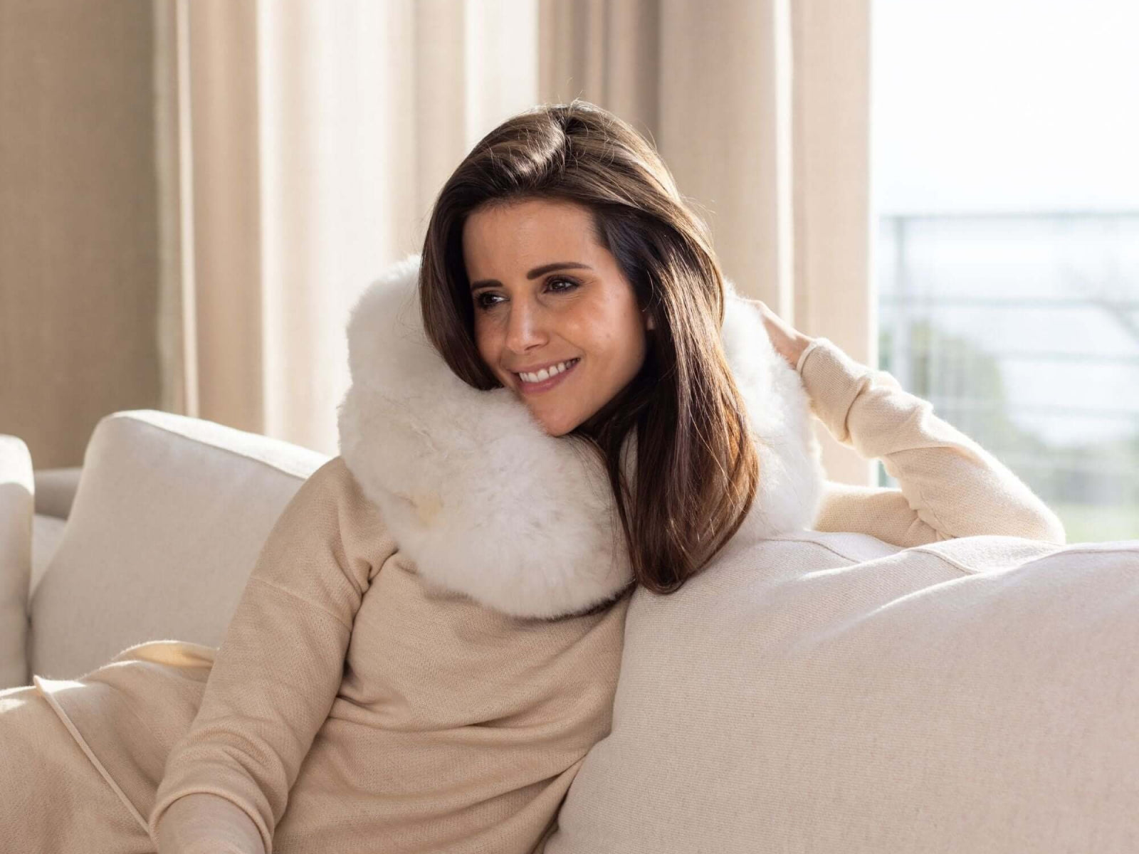 Woman lounging on a sofa with a white plush travel pillow wrapped around her neck.
