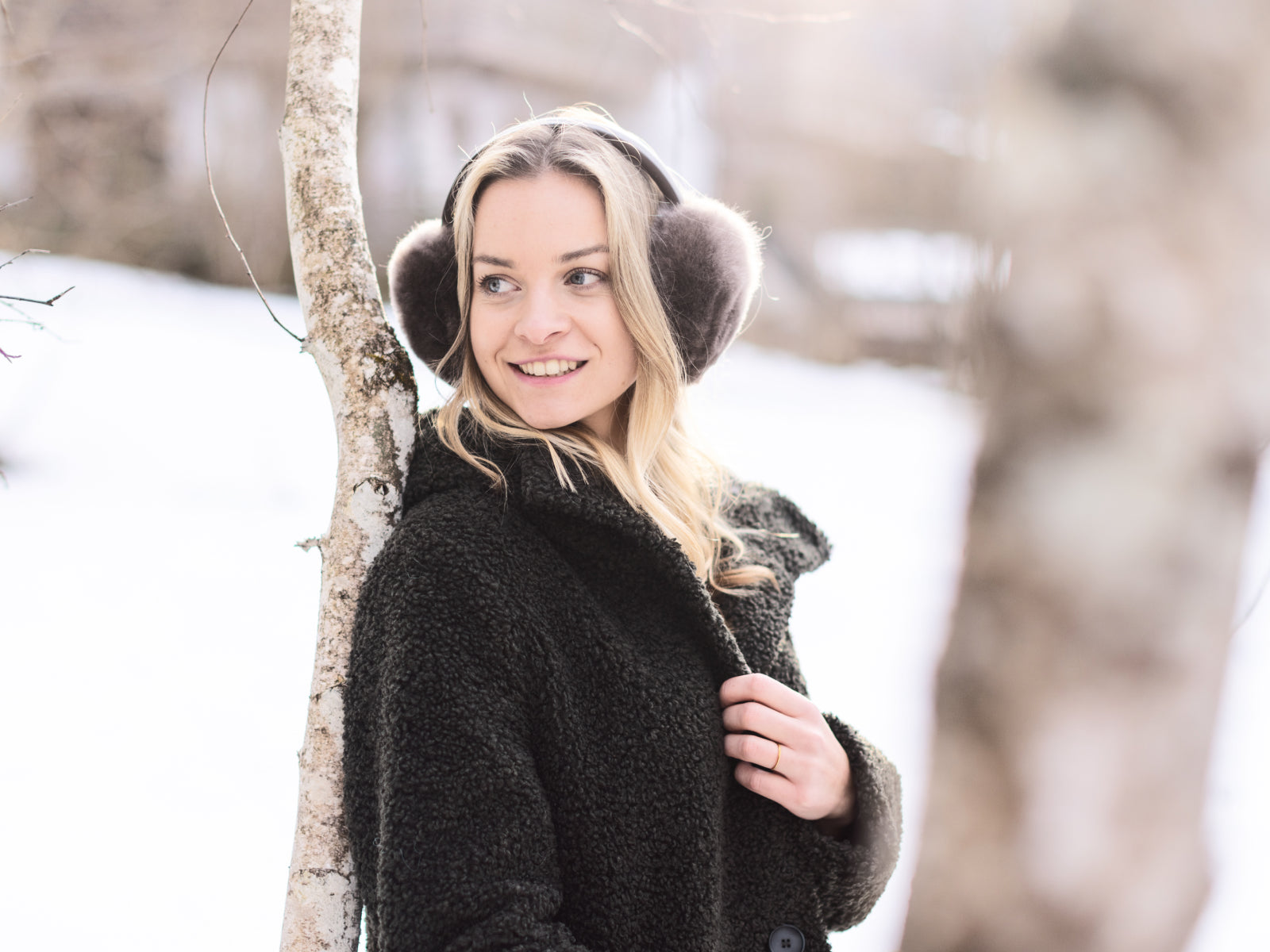 Woman outdoors wearing dark fluffy OLMO ear warmers with a black teddy coat in a winter scene