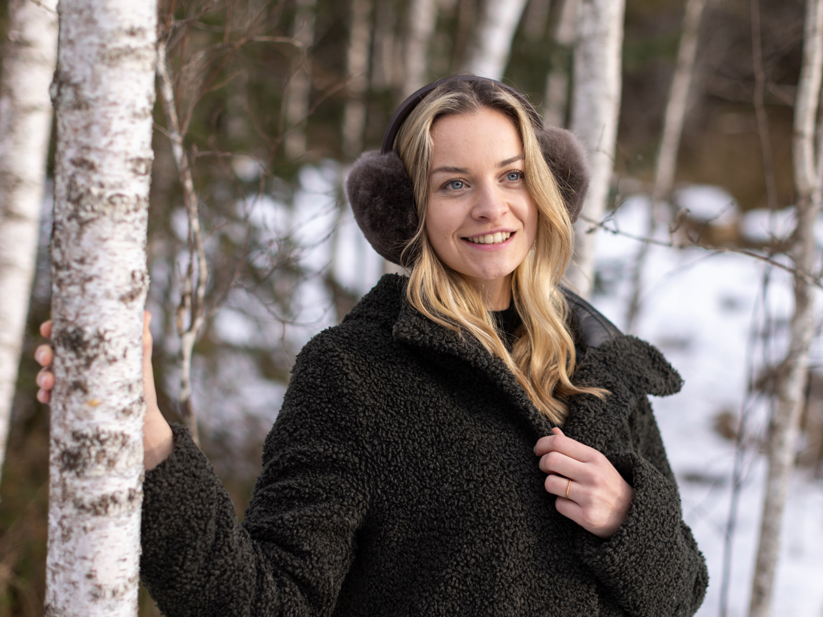 Smiling woman in a black teddy coat wearing dark brown fluffy earmuffs with a headband in a snowy forest