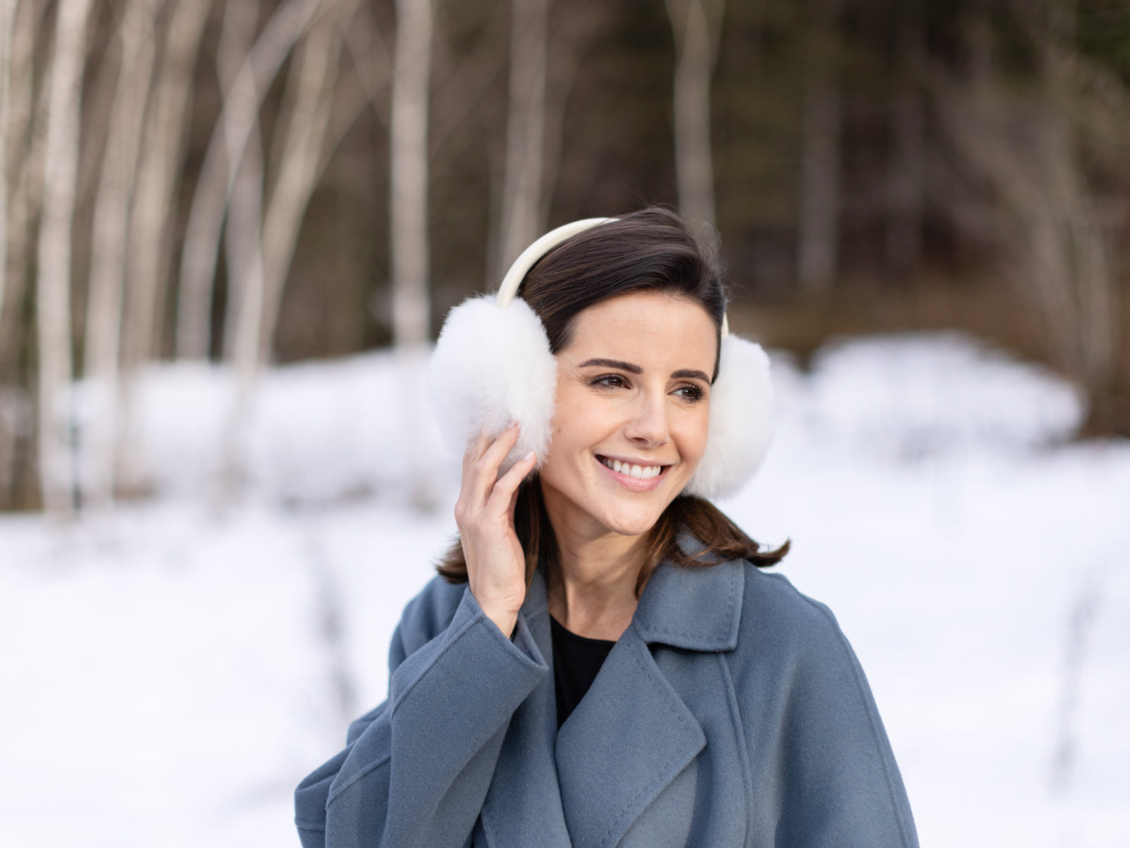 Smiling woman in a blue coat wearing white fluffy ear muffs in a snowy woodland.