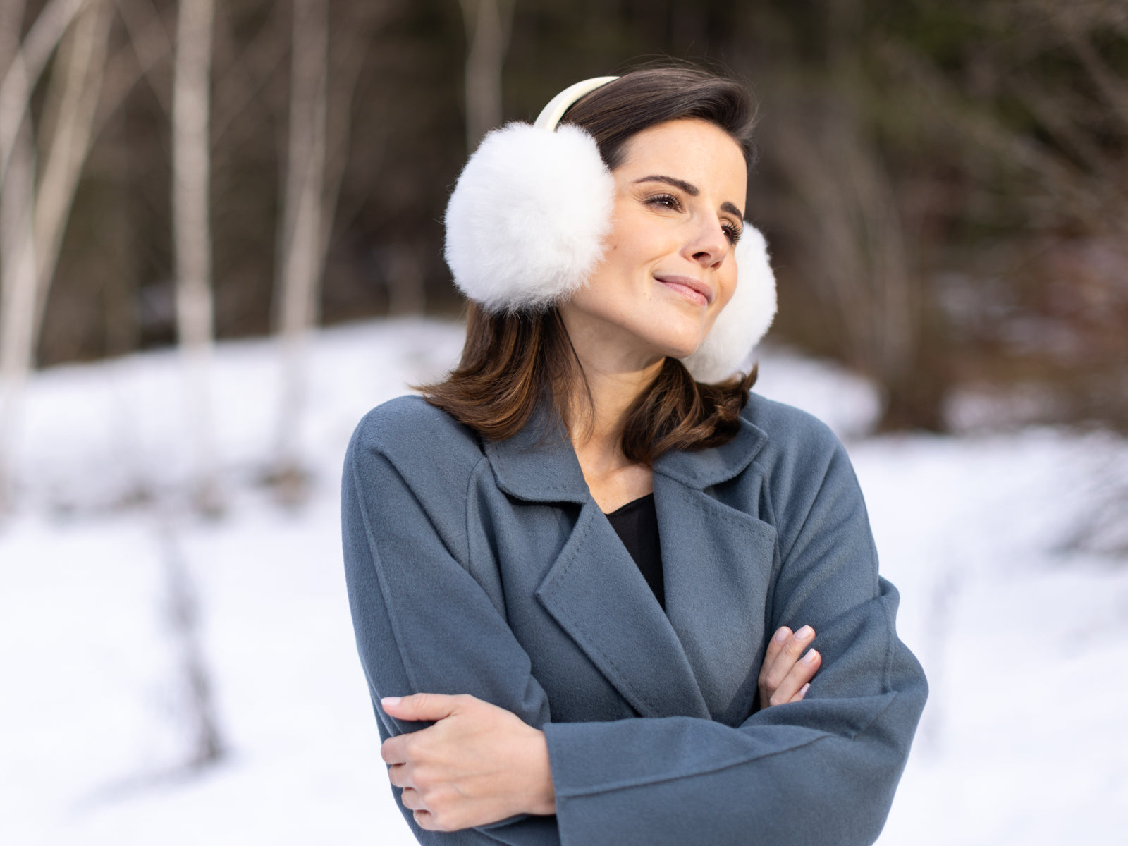 Woman wearing white fluffy ear warmers with a bluish gray coat standing among birch trees