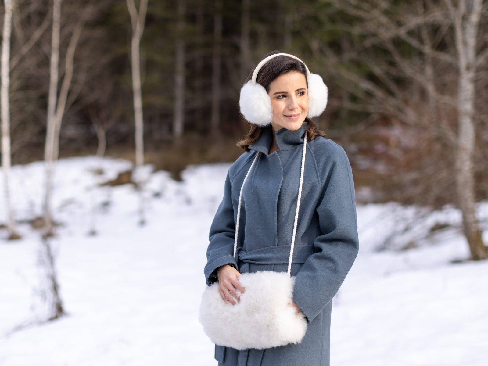 Woman in a blue coat wearing white fluffy ear warmers with a matching fluffy crossbody bag in a snowy landscape