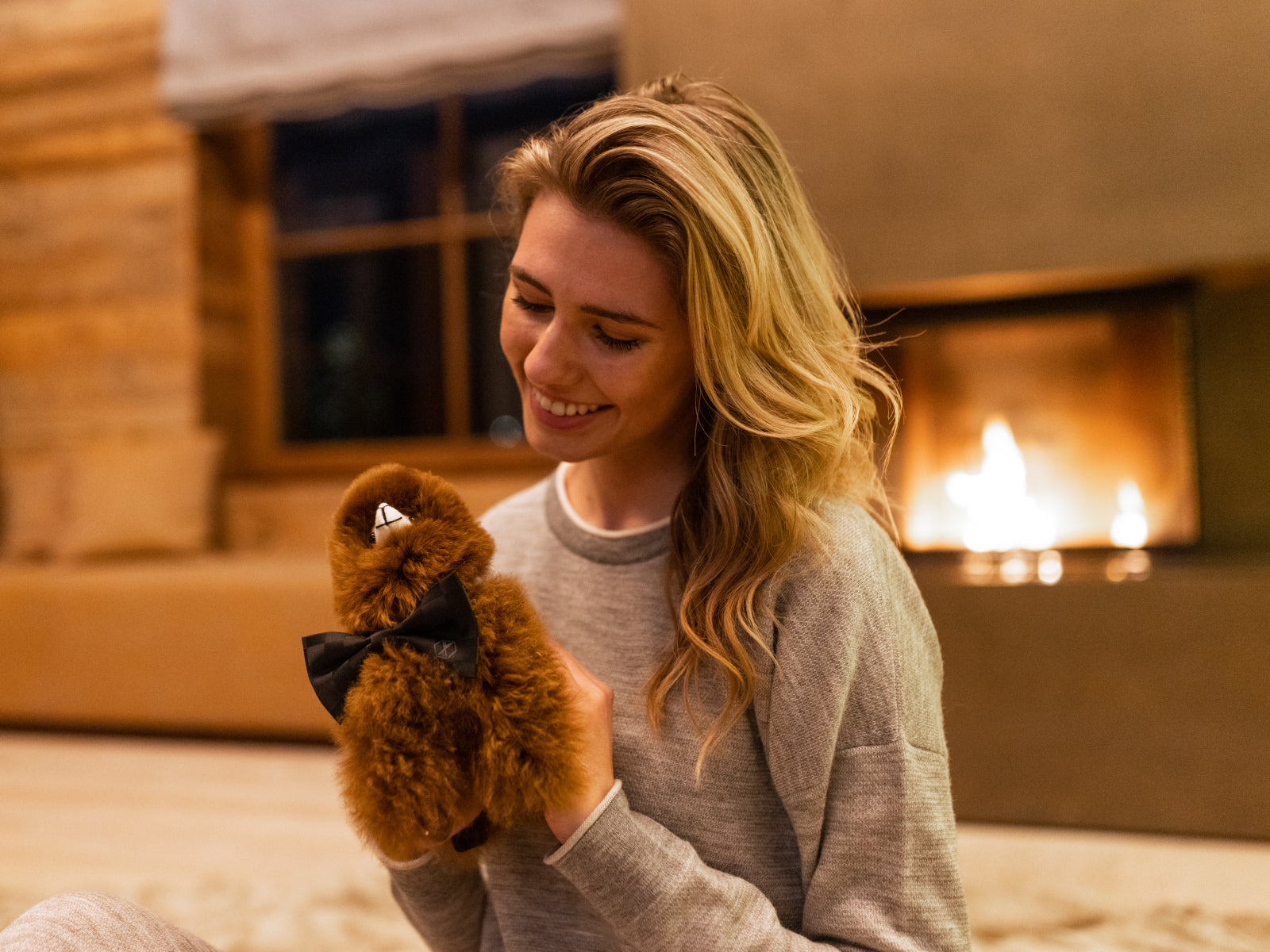 Smiling woman cuddling the brown Alpaca Paco plush with a bow tie in a cozy living room