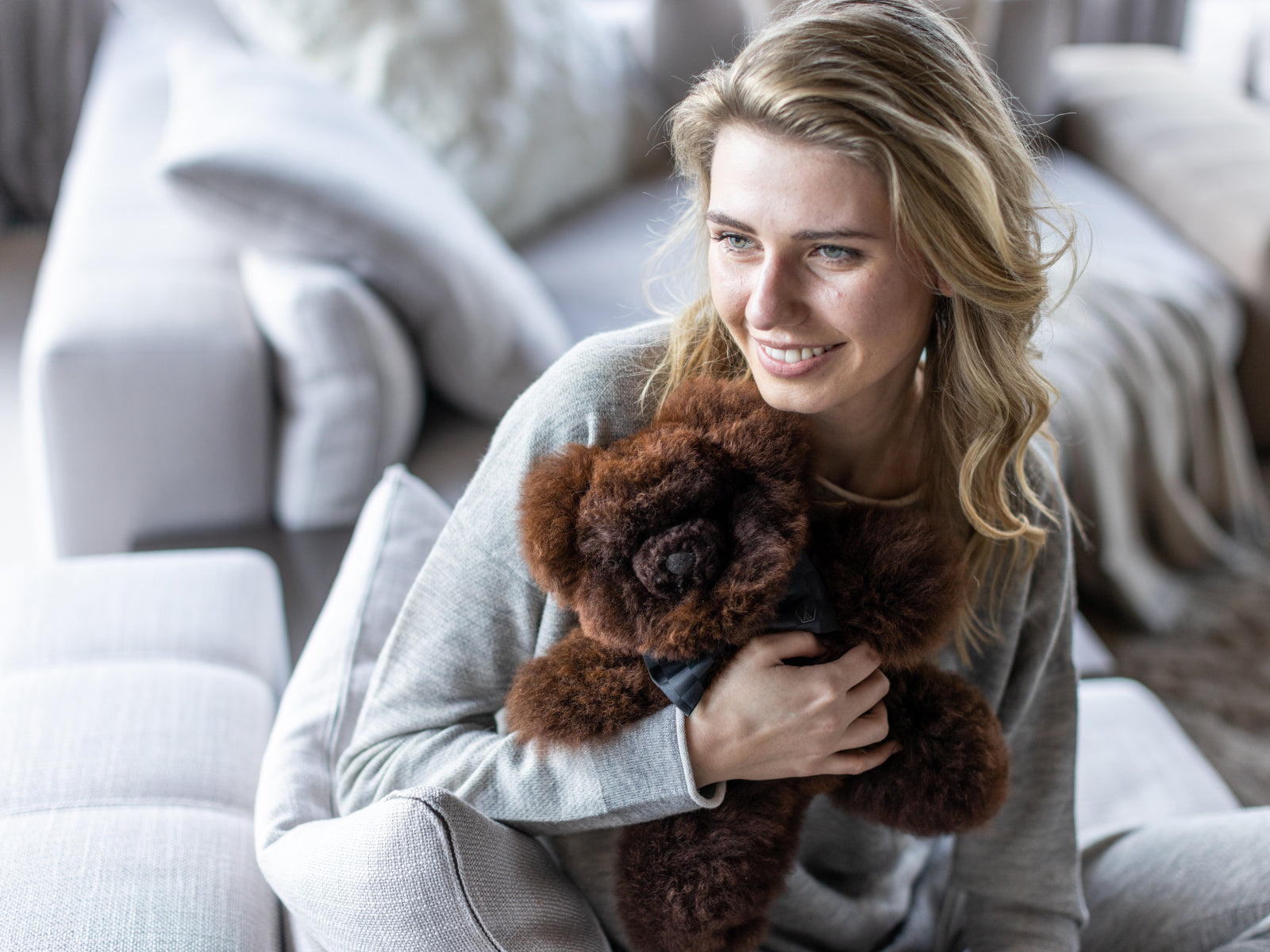 Woman on a gray sofa cuddling a fluffy brown teddy bear with a black bow tie.