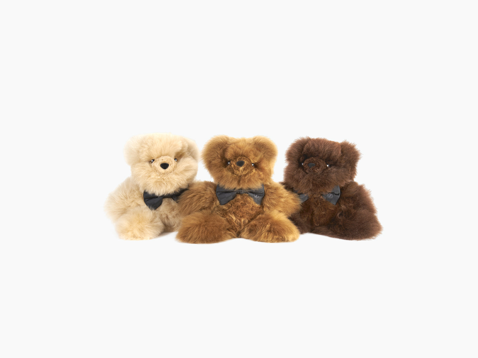 Three plush teddy bears in beige, light brown, and dark brown with black bow ties placed together.