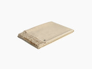 Beige folded throw blanket with fringed edges on a white studio backdrop.