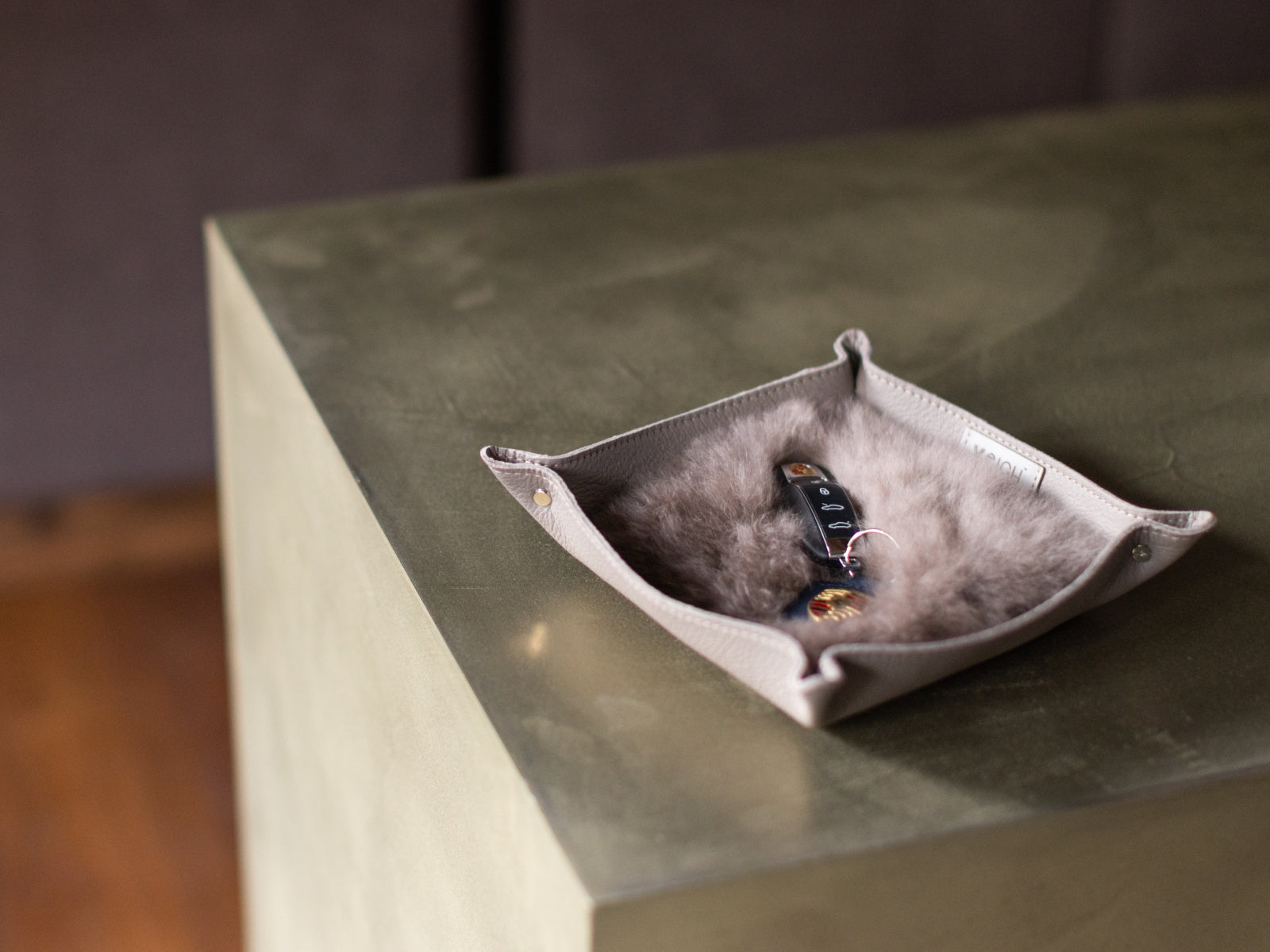 Beige leather catchall tray lined with grey fur sits on a metal surface with keys inside.