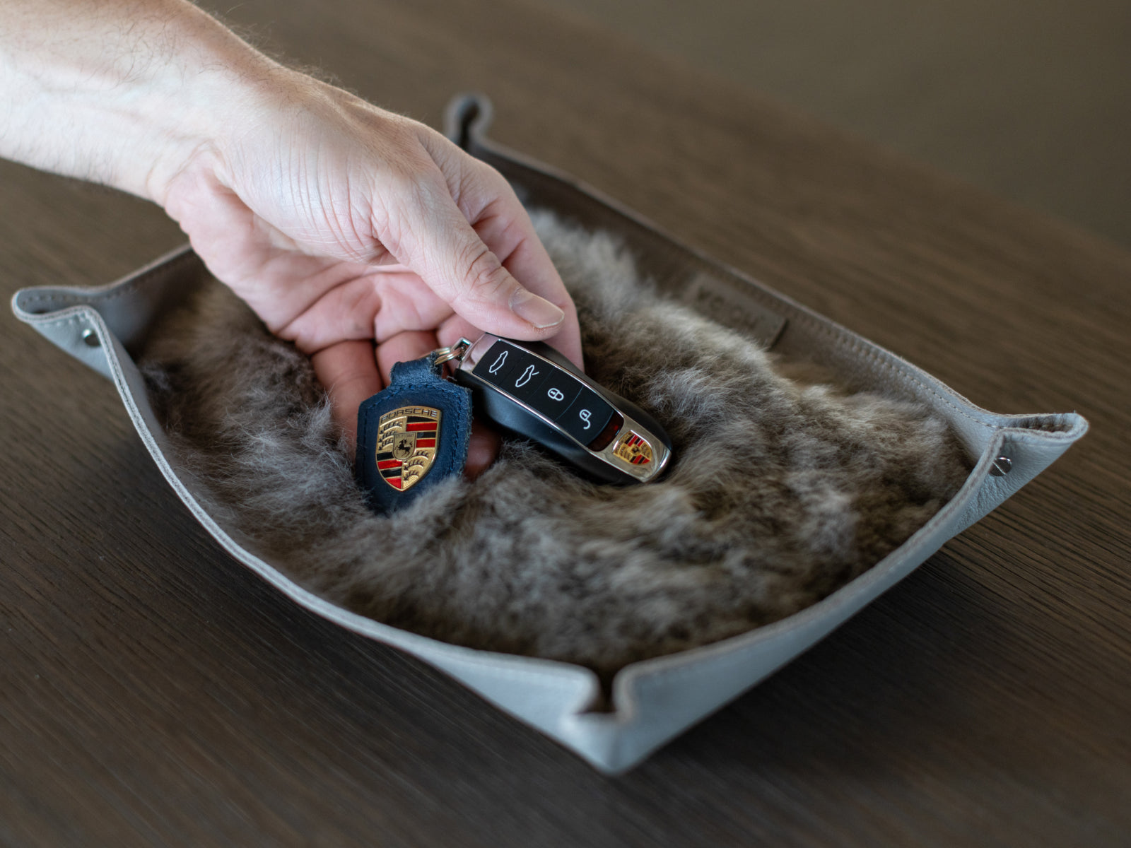 Hand placing a Porsche key fob and badge into a light gray leather catchall tray with plush fur lining