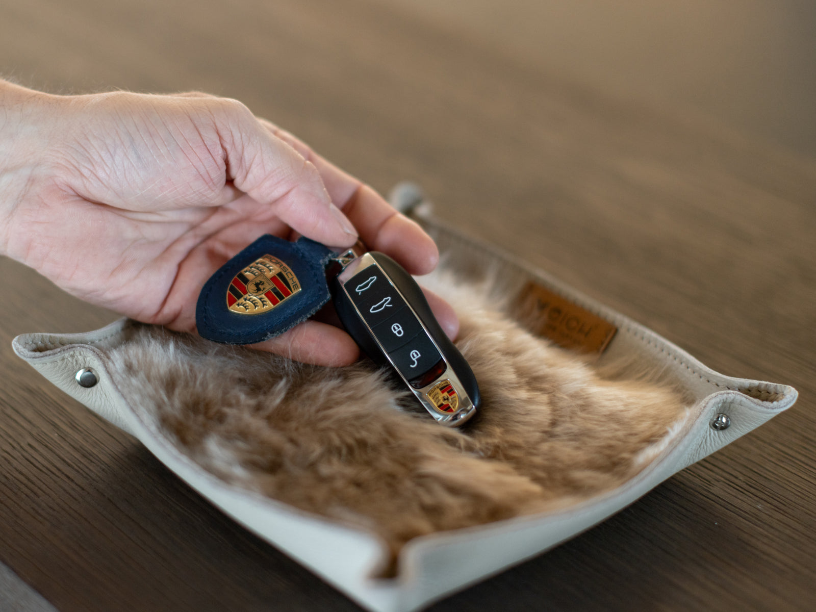 Hand holds a Porsche key fob over a white fur lined WEICH catchall tray.