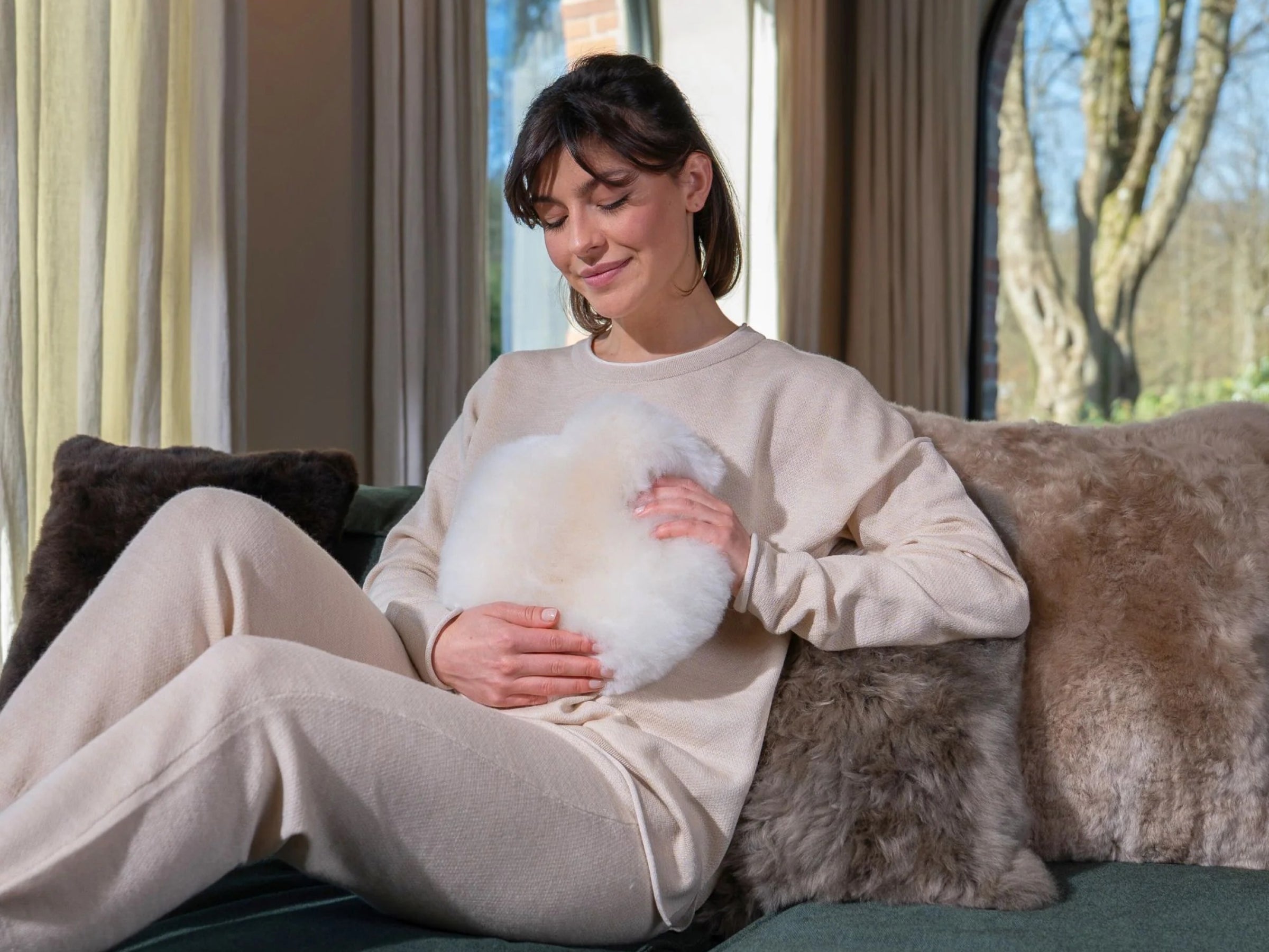 Woman in beige loungewear sits on a sofa cuddling a soft white alpaca fiber heart shaped hot water bottle.